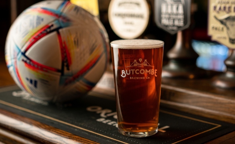 a glass of butcombe beer/cider on a table, next to a rugby ball and a screen playing a rugy match